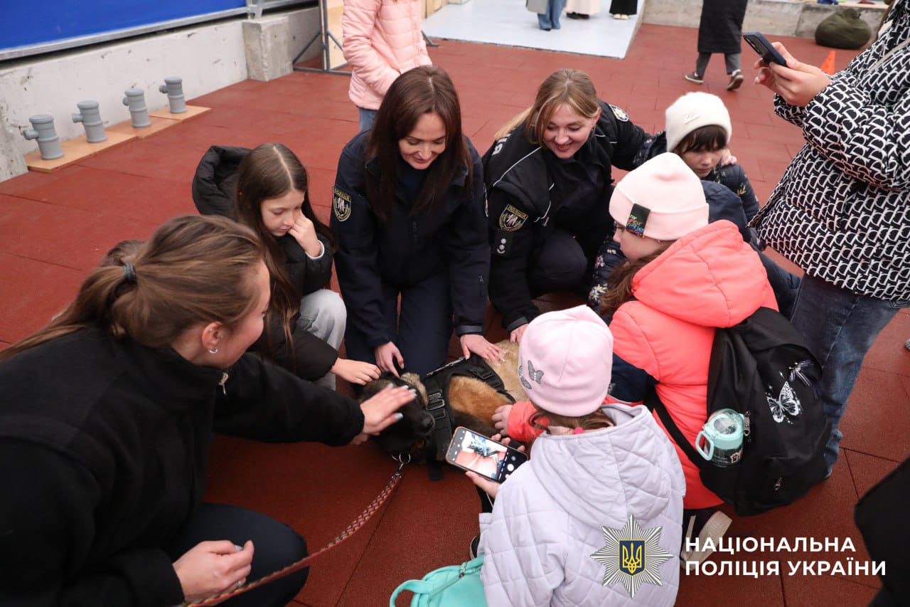 Діти та поліцейські гладять службового собаку, який лежить на підлозі.