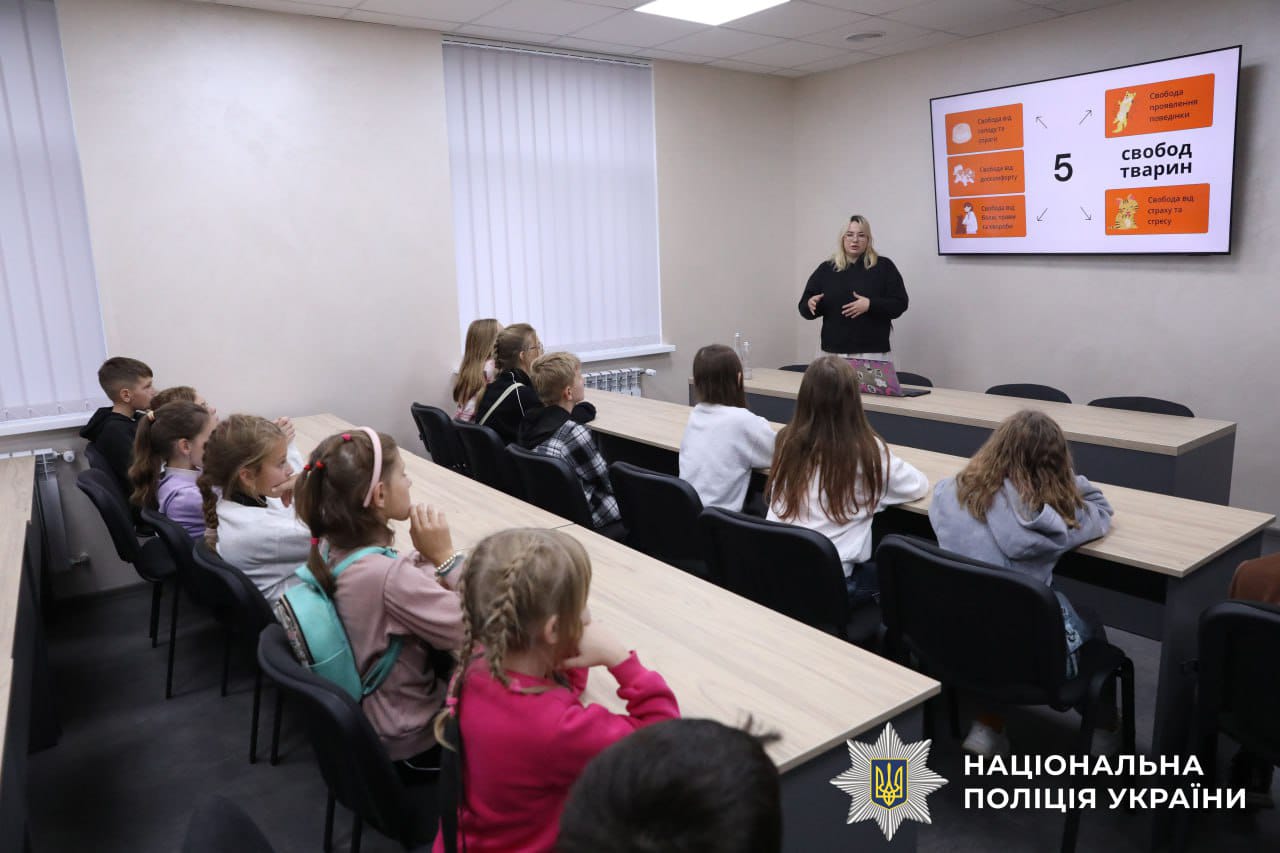 Діти сидять за столами і слухають лекцію. Поліцейська розповідає інформацію, стоячи перед учнями.