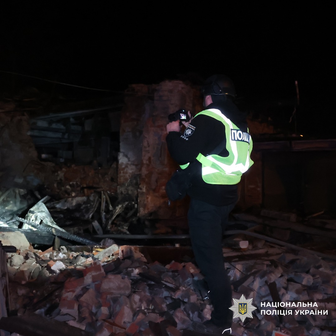 Поліцейський у бронежилеті та світловідбивному жилеті фотографує руйнування будівлі вночі після обстрілу. На передньому плані уламки цегли й зруйновані конструкції.