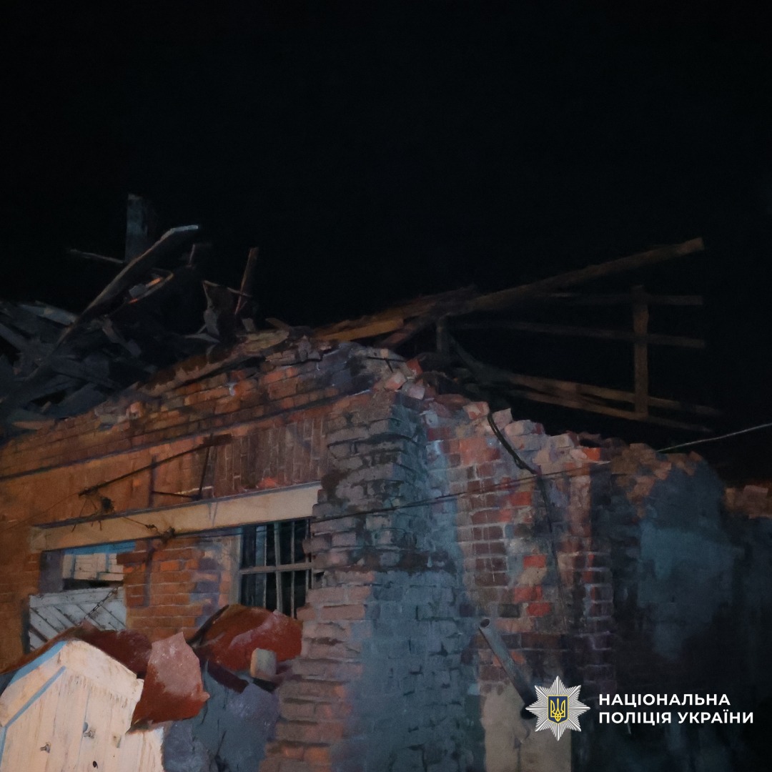 Нічне фото зруйнованої цегляної будівлі з частково обваленим дахом і стінами, серед уламків та дерев’яних балок. Освітлення штучне, ймовірно, від прожекторів, що підкреслює масштаби руйнування.