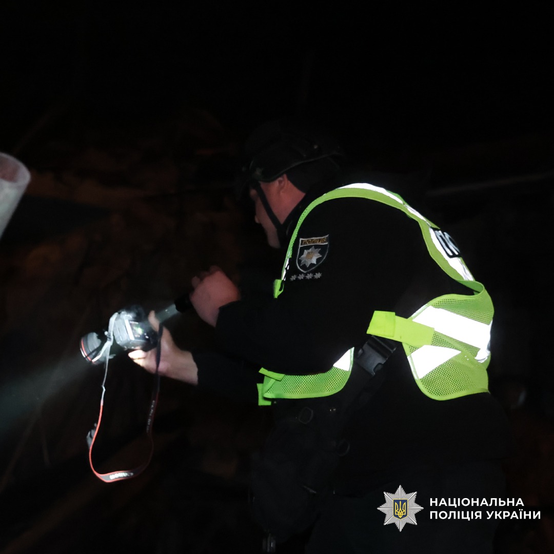 Поліцейський у бронежилеті та світловідбивному жилеті фотографує руйнування будівлі вночі після обстрілу. На передньому плані уламки цегли й зруйновані конструкції.