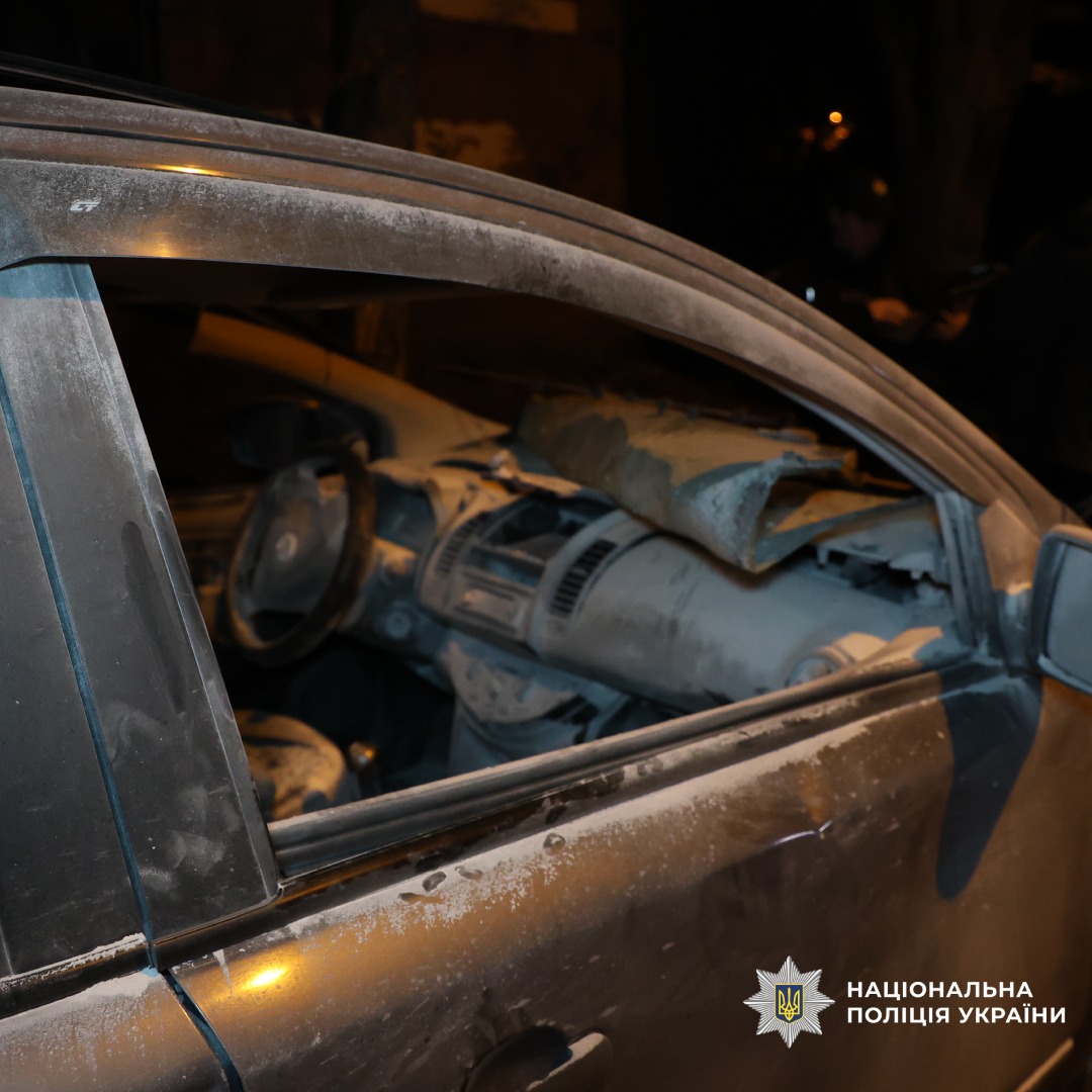 Пошкоджений автомобіль, зафіксований через водійське вікно. Салон вкритий товстим шаром пилу та уламків, на панелі великий обломок, лобове скло відсутнє або розбите. Рульове колесо та сидіння також забруднені. Темна атмосфера вказує на нічний час або слабке освітлення. У тлі нечіткі постаті та стіна будівлі.