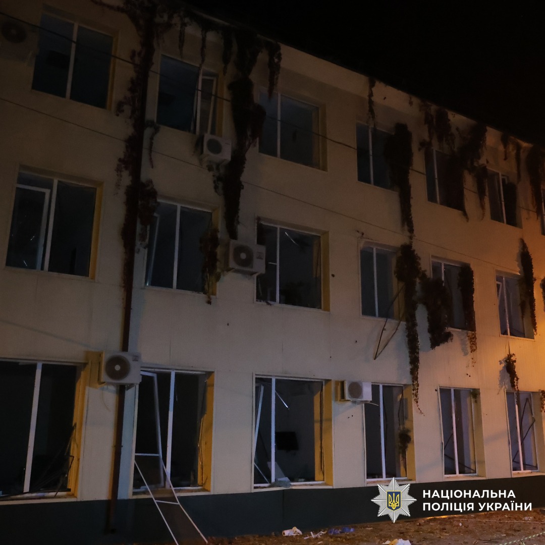 Пошкоджена багатоповерхова будівля вночі, з вибитими вікнами та звисаючими кондиціонерами. Штучне освітлення підкреслює сліди руйнування.