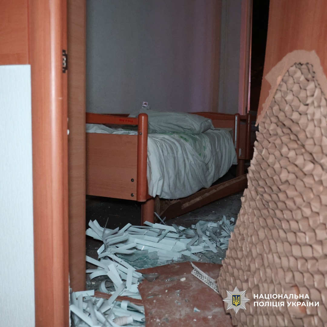 Пошкоджене приміщення лікарні: дерев’яне ліжко з білою постільною білизною на тлі, розбиті двері. Праворуч розбиті двері.