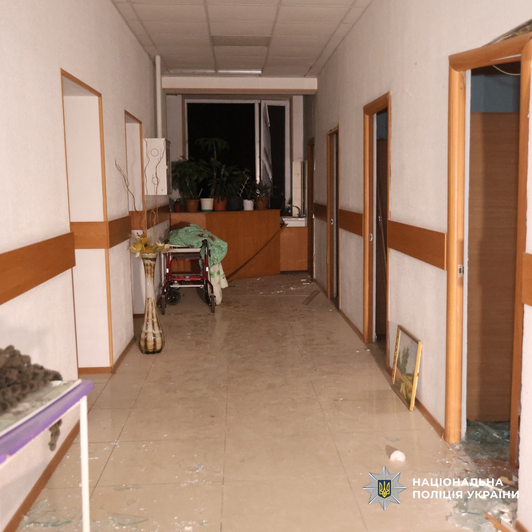 Пошкоджений коридор лікарні: на підлозі розбите скло та уламки, відкриті двері ведуть у порожні кімнати. Видно інвалідний візок і медичне обладнання. Наприкінці коридору пост медсестри з вазонами та столом. Атмосфера тривожна.