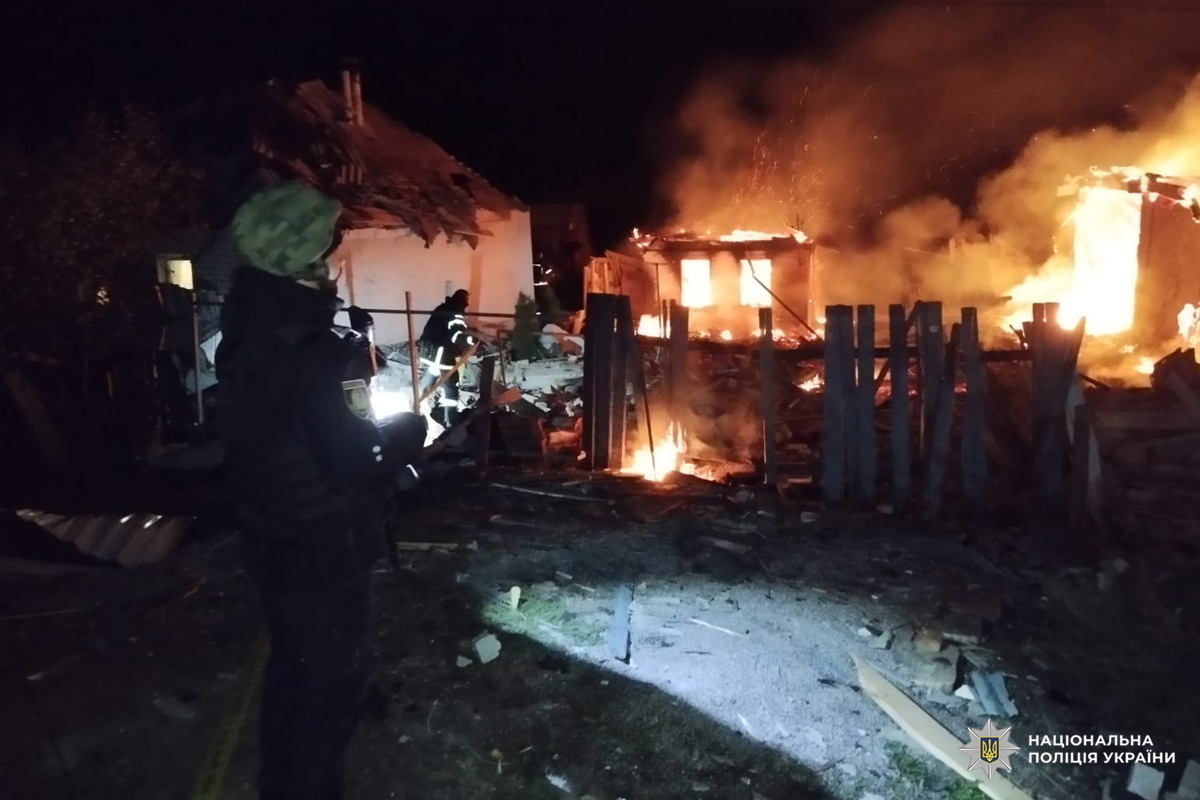 Нічна сцена пожежі: зруйнована будівля охоплена полум’ям. На передньому плані поліцейський, тримає ліхтарик і пристрій зв’язку. На тлі інші екстрені служби працюють серед вогню та уламків.