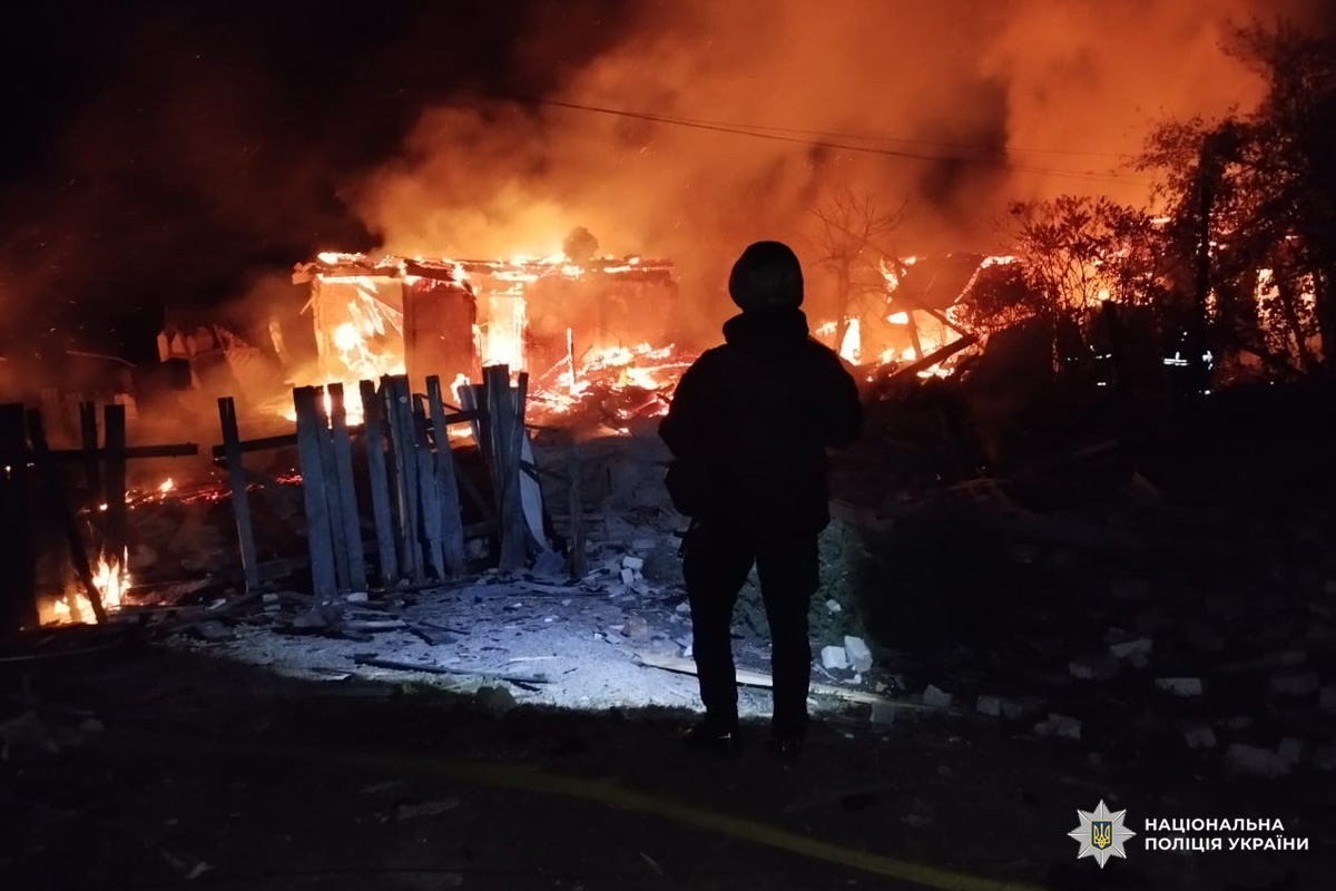 Велика пожежа охоплює житловий район уночі. На передньому плані силует поліцейського, який спостерігає за полум’ям. Вогонь знищив кілька споруд, дим і язики полум’я здіймаються високо в небо. На землі уламки та пошкоджені огорожі.