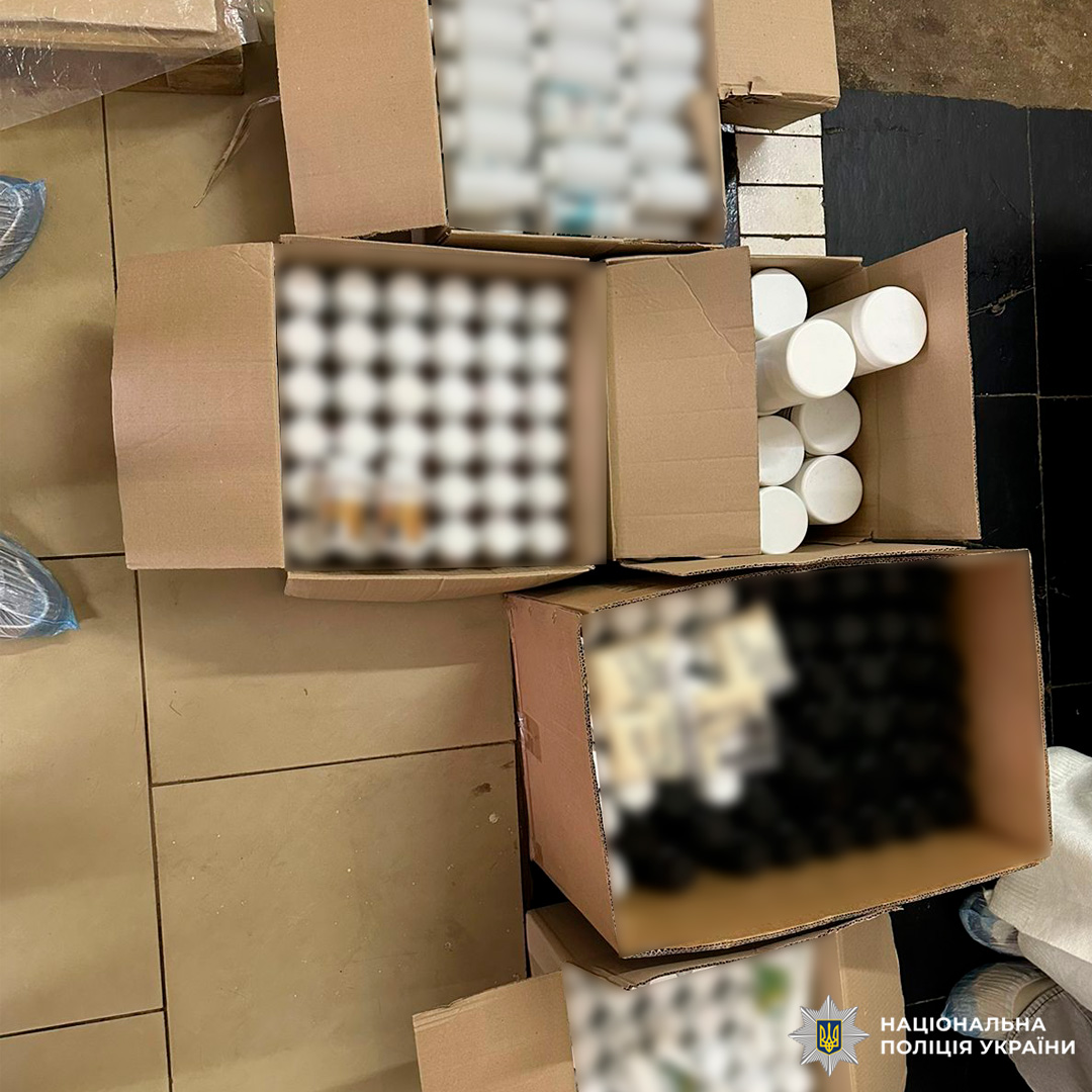 Зображено чотири відкриті картонні коробки, розміщені на плитковій підлозі. У коробках різні медичні товари: білі флакони, чорні та білі циліндричні контейнери, упаковані предмети.