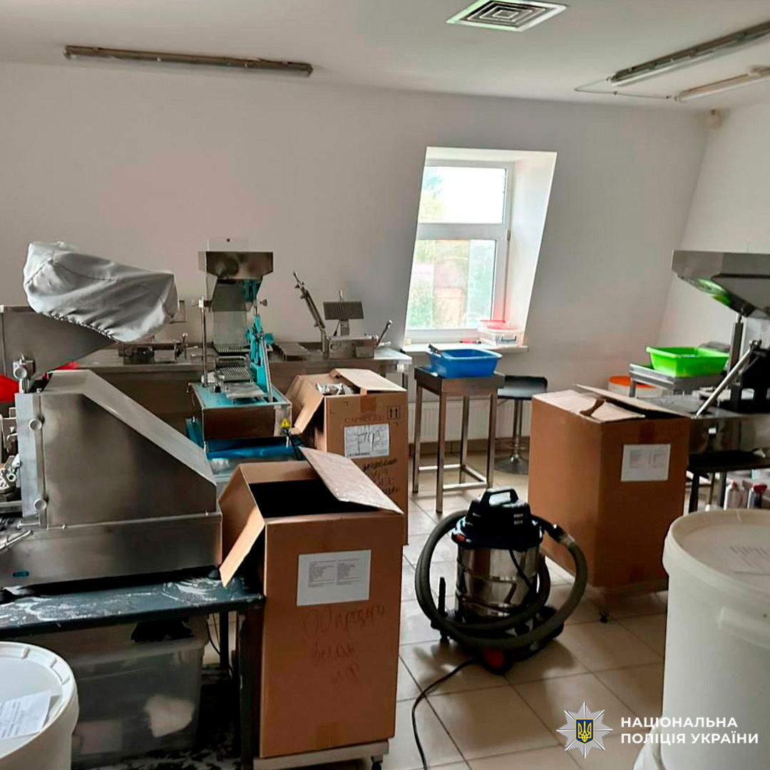 На зображенні виробниче приміщення з металевими машинами, картонними коробками, пластиковими контейнерами, пилососом і меблями. Підлога плиткова, стіни білі.