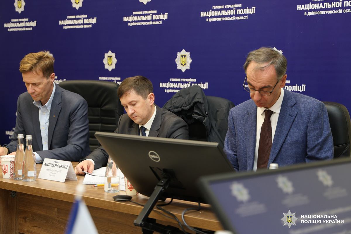 Троє чоловіків у діловому одязі сидять за конференційним столом під час офіційної зустрічі. На столі документи, пляшки з водою та чашки з кавою. Один із чоловіків працює за ноутбуком. На фоні банер з написом  'Головне управління Національної поліції в Дніпропетровській області'.