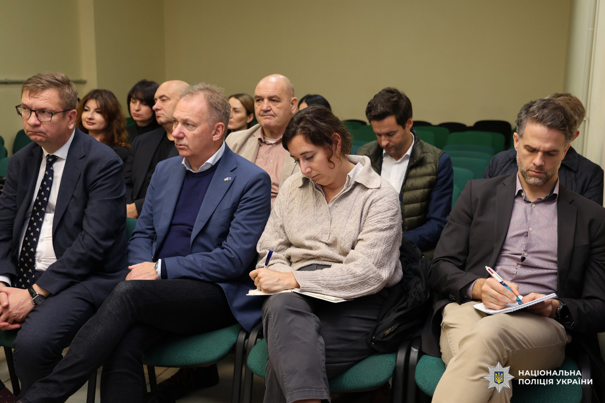 Група людей сидить у кімнаті під час офіційної зустрічі. Деякі учасники роблять нотатки. У приміщенні зелені стільці та нейтральні стіни.