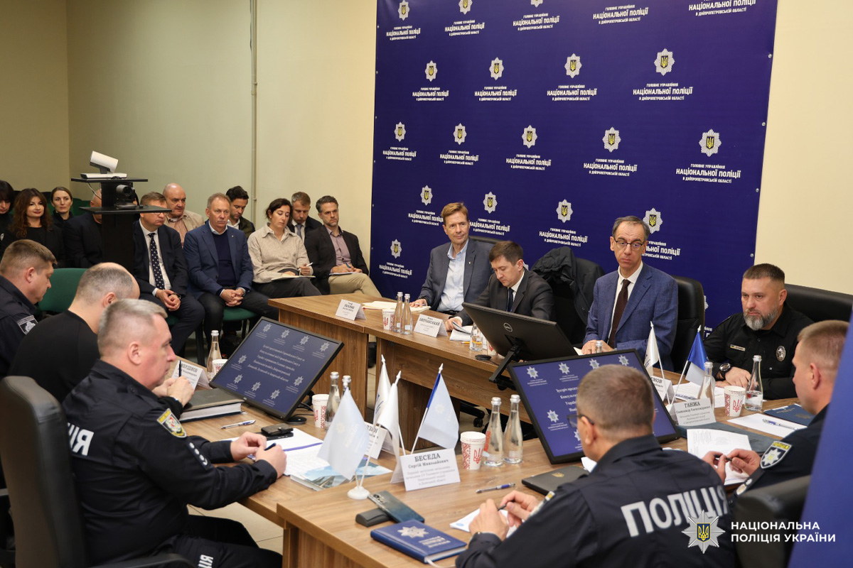 Офіційна зустріч представників Національної поліції України та ЄС. Учасники  у формі та цивільному одязі сидять за великим столом із мікрофонами, табличками з іменами, документами та цифровими екранами. На фоні  логотип і назва Національної поліції України.