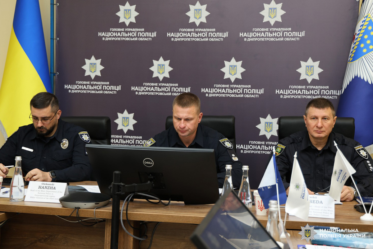 Троє поліцейських у формі сидять за столом під час офіційної зустрічі або пресконференції. Перед ними мікрофони, ноутбуки та документи.