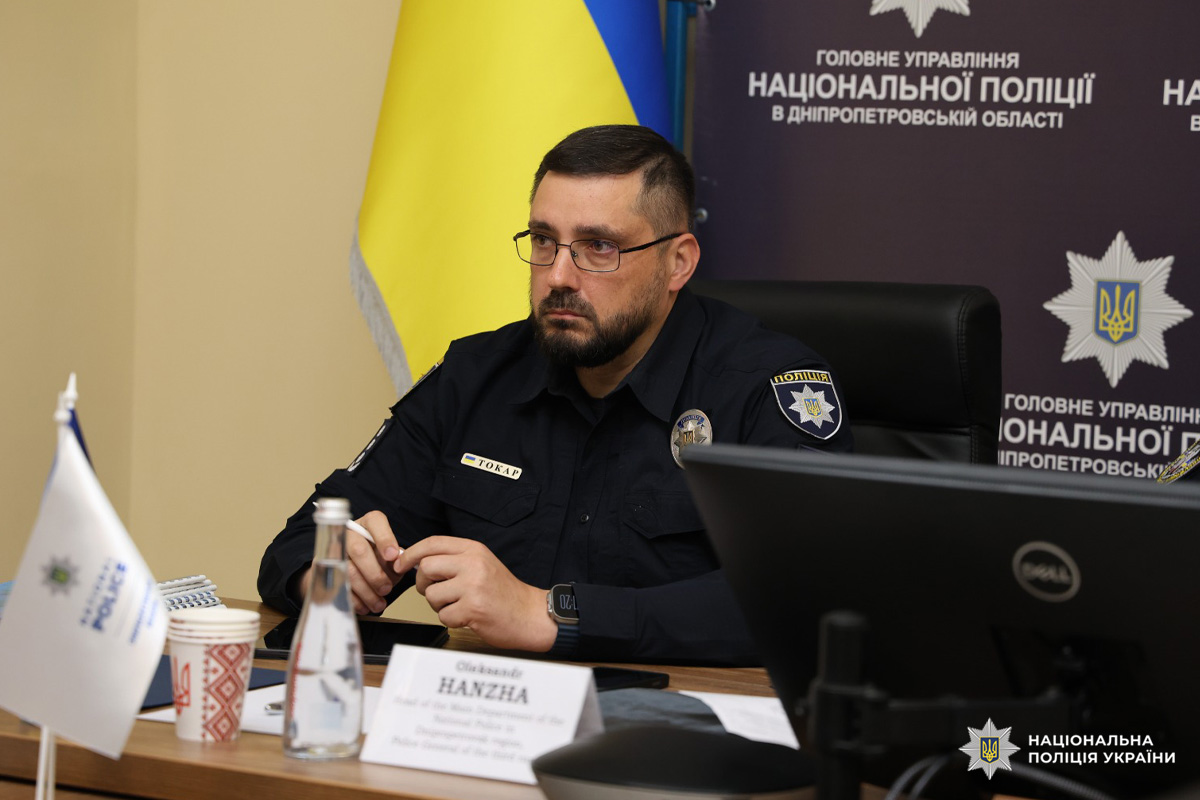 Чоловік у поліцейській формі сидить за столом під час офіційної зустрічі або пресконференції. На столі пляшка води, чашка кави, монітор.