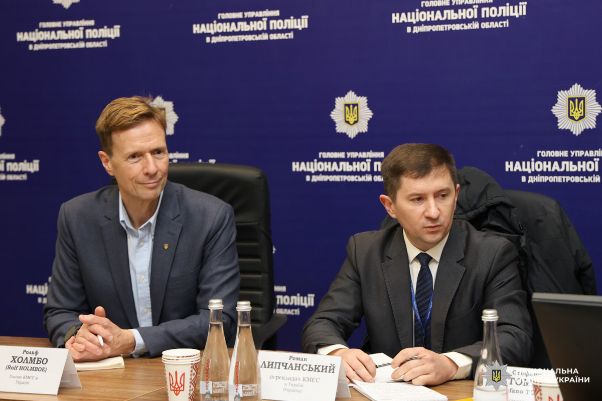 Двоє чоловіків сидять за конференційним столом під час офіційної зустрічі. На фоні емблема та напис 'Головне управління Національної поліції в Дніпропетровській області'. Один із чоловіків у світлій сорочці та синьому піджаку, інший у темному костюмі з краваткою.