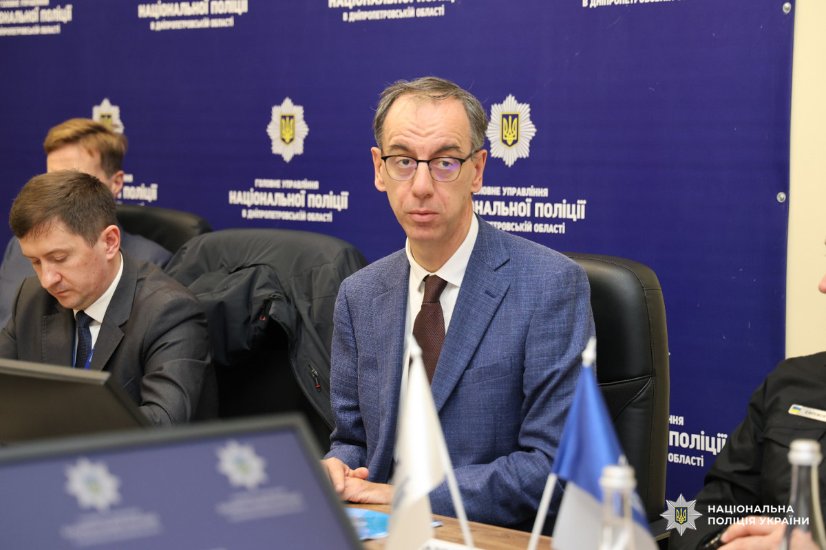 Група осіб сидить за конференційним столом під час офіційної зустрічі або брифінгу. На фоні синій банер з емблемою та написами 'Національна поліція у Дніпропетровській області'. Чоловік у центрі в синьому костюмі та окулярах. На столі ноутбуки, документи, прапори.
