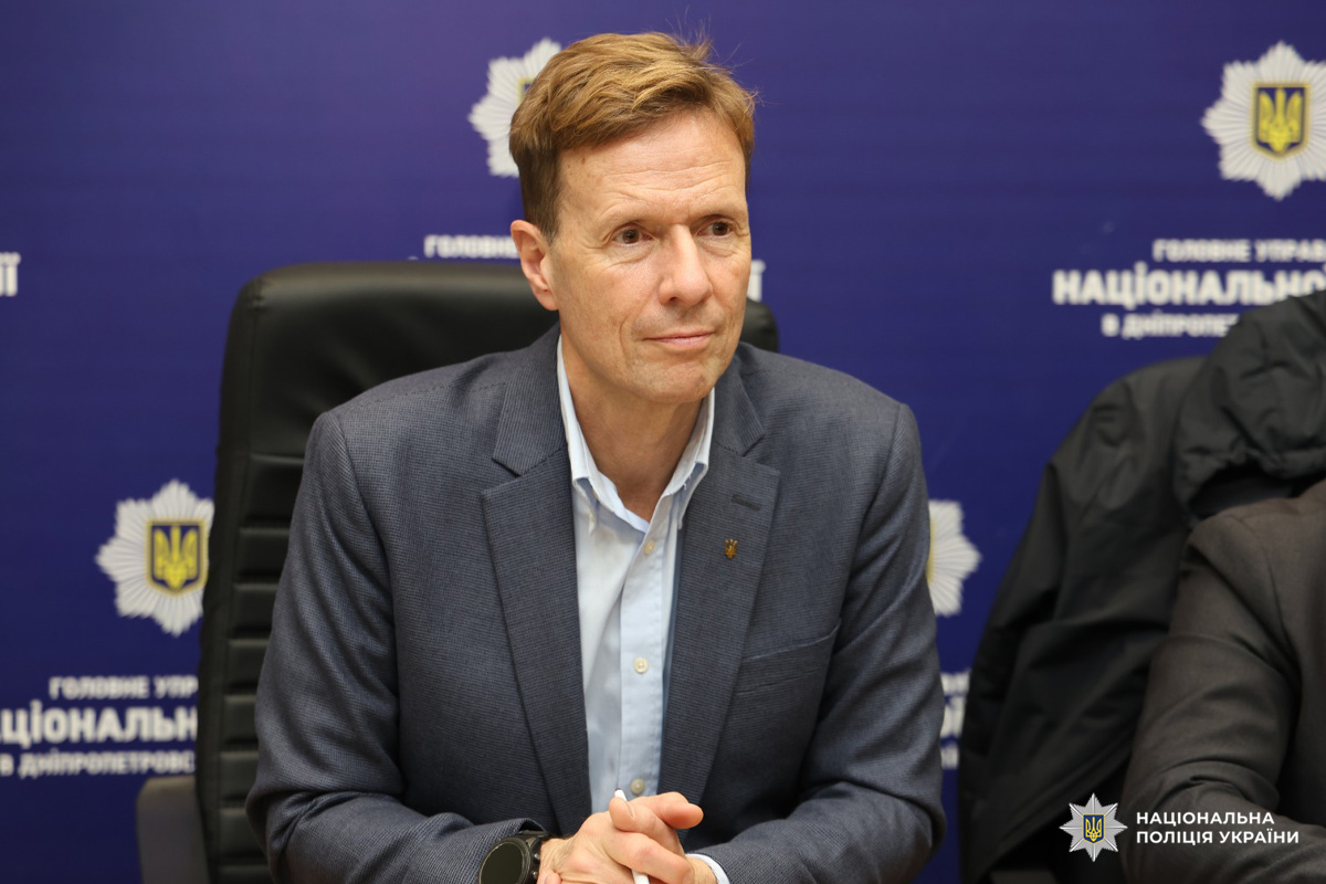 Чоловік у темному піджаку та світлій сорочці сидить за столом на фоні синього банера.Атмосфера свідчить про офіційну зустріч або пресконференцію, ймовірно, пов’язану з правоохоронною діяльністю.