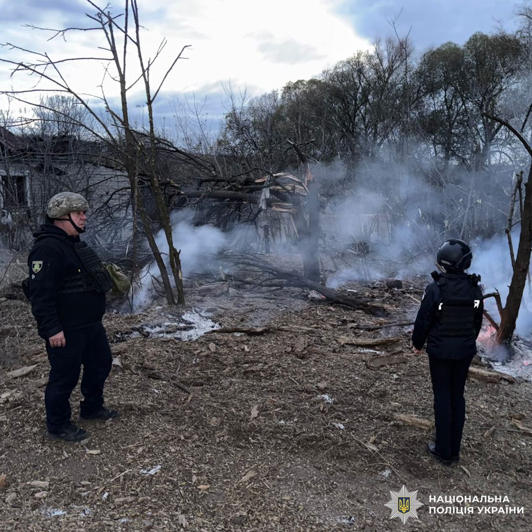 Двоє поліцейських стоять біля задимленої ділянки на відкритій місцевості з пошкодженою будівлею на тлі. На землі сліди пожежі або вибуху, серед дерев без листя.
