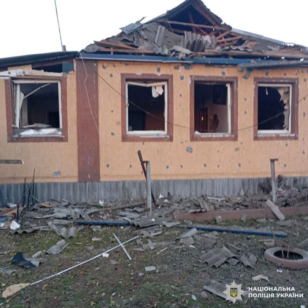 Зруйнований житловий будинок із частково обваленим дахом, вибитими вікнами та слідами ураження на стінах, ймовірно від уламків або обстрілу. На передньому плані розкидані уламки дерева, металу та автомобільні шини.