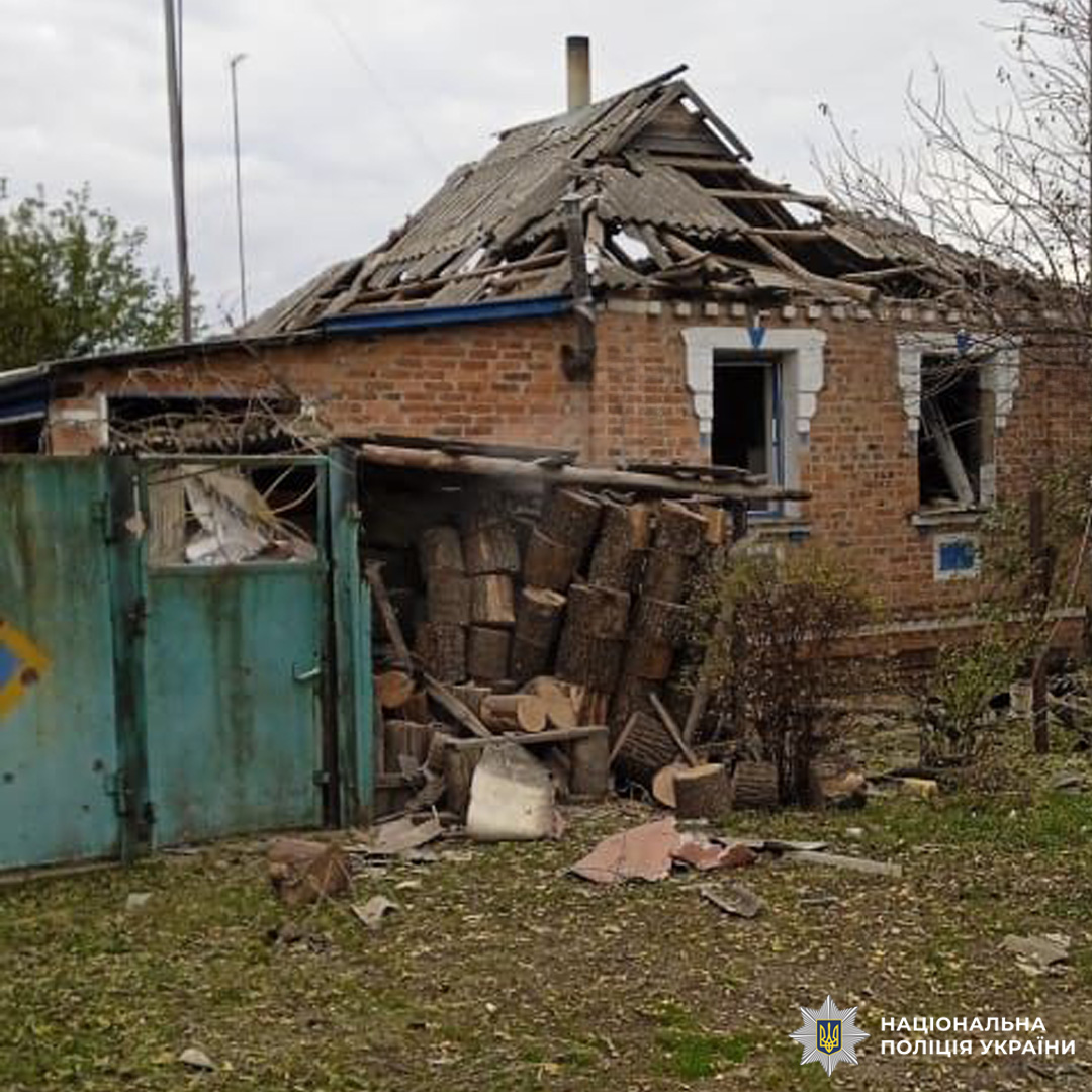 Цегляний будинок із частково обваленим дахом, пошкодженими вікнами та відкритим інтер’єром. Перед фасадом вертикально складені дрова, навколо уламки цегли, дерева та інших матеріалів.