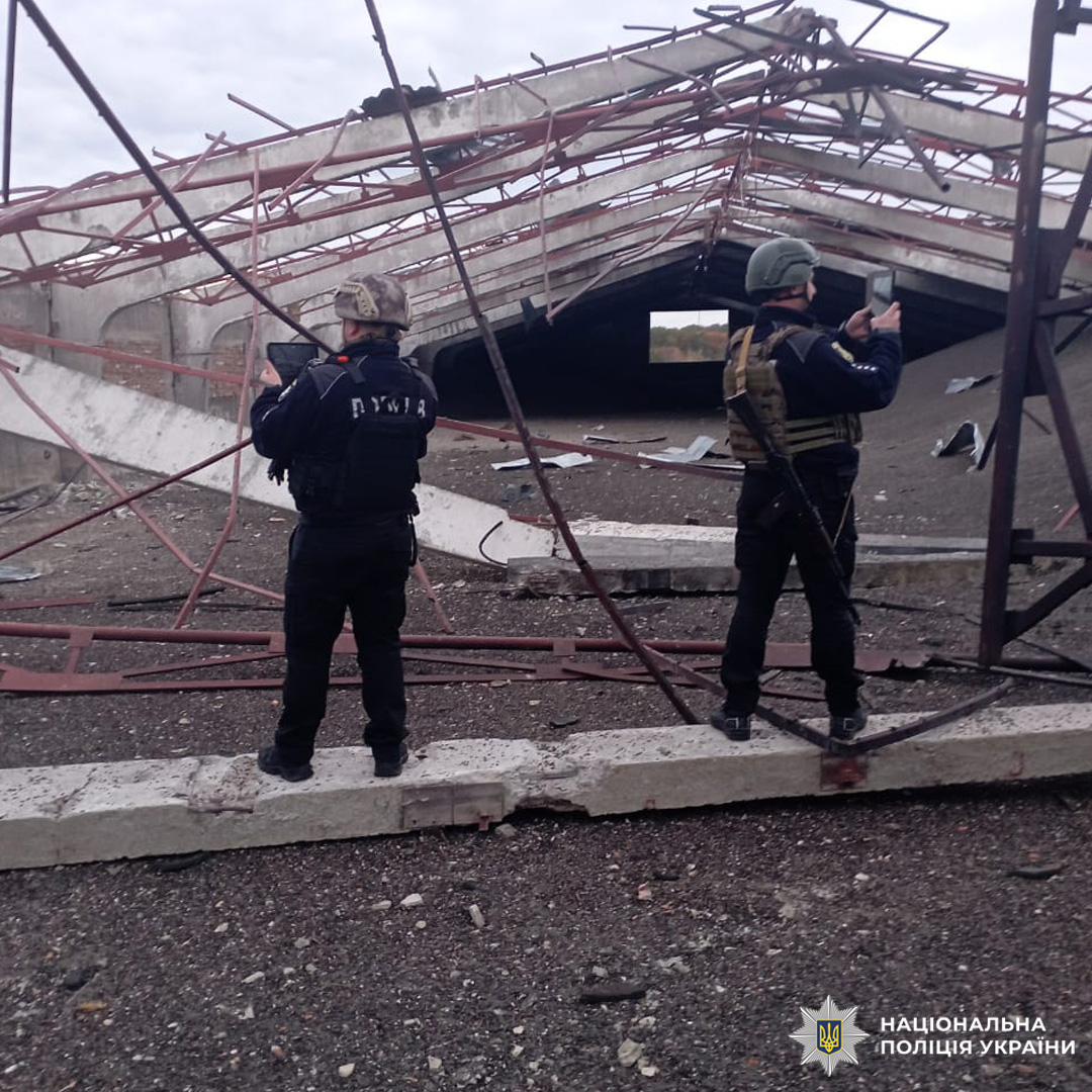 Двоє правоохоронців стоять перед зруйнованою будівлею з обваленим дахом і металевими конструкціями. На землі — уламки, небо похмуре.