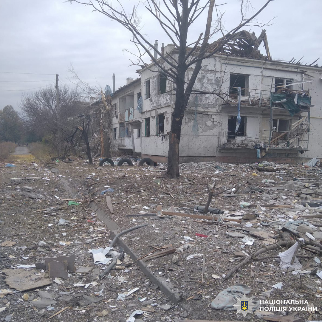 Зруйнована будівля з обваленим дахом і розбитими вікнами стоїть у спустошеній місцевості. Перед нею голе дерево, навколо розкидані уламки дерева, металу та паперу.