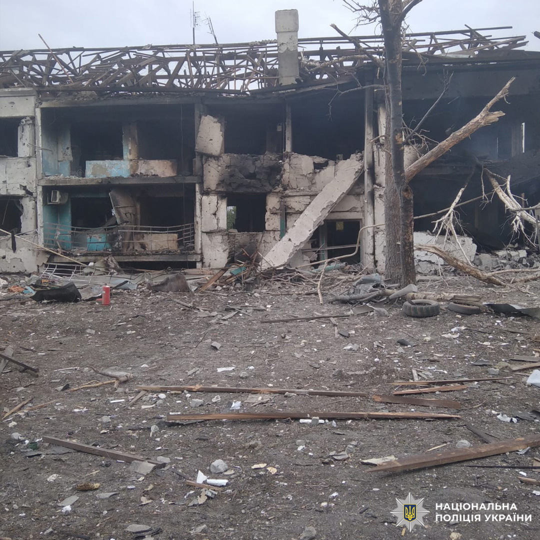 Багатоповерхова будівля з серйозними пошкодженнями фасаду та внутрішніх конструкцій, імовірно внаслідок вибуху або бойових дій. Частина даху обвалилася, стіни обгорілі та зруйновані. На передньому плані уламки бетону, дерева й металу, а також пошкоджене безлисте дерево з обламаними гілками.
