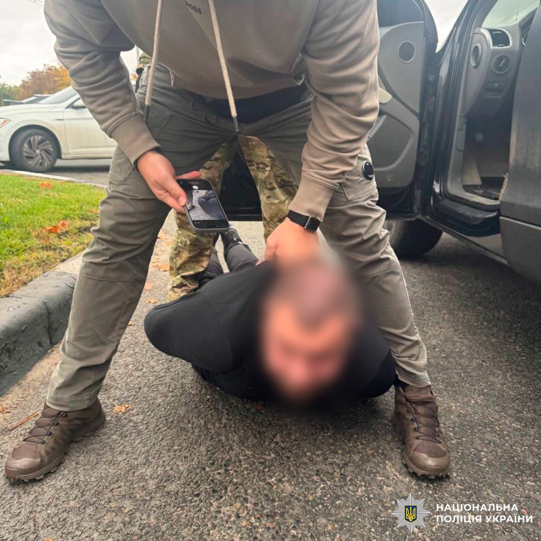 Особа лежить обличчям донизу на асфальті з руками, зафіксованими за спиною. Правоохоронці в тактичному спорядженні здійснюють затримання; один із них тримає смартфон, ймовірно для фіксації події. Поруч службовий автомобіль із відкритими дверима.