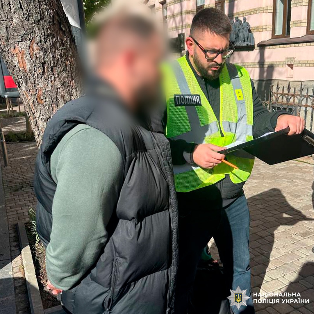 Правоохоронець проводить документування особи з розмитим обличчям. Сцена відбувається на асфальтованій ділянці біля будівлі.