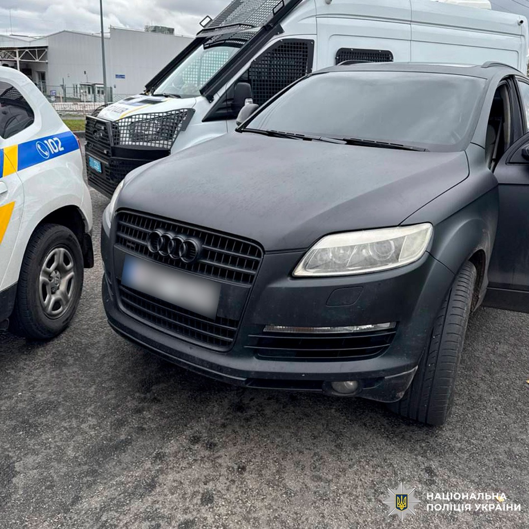 Чорний позашляховик Audi зупинений між поліцейським автомобілем з номером "102" та білим фургоном із захисною сіткою на лобовому склі. Водійські двері Audi відкриті.