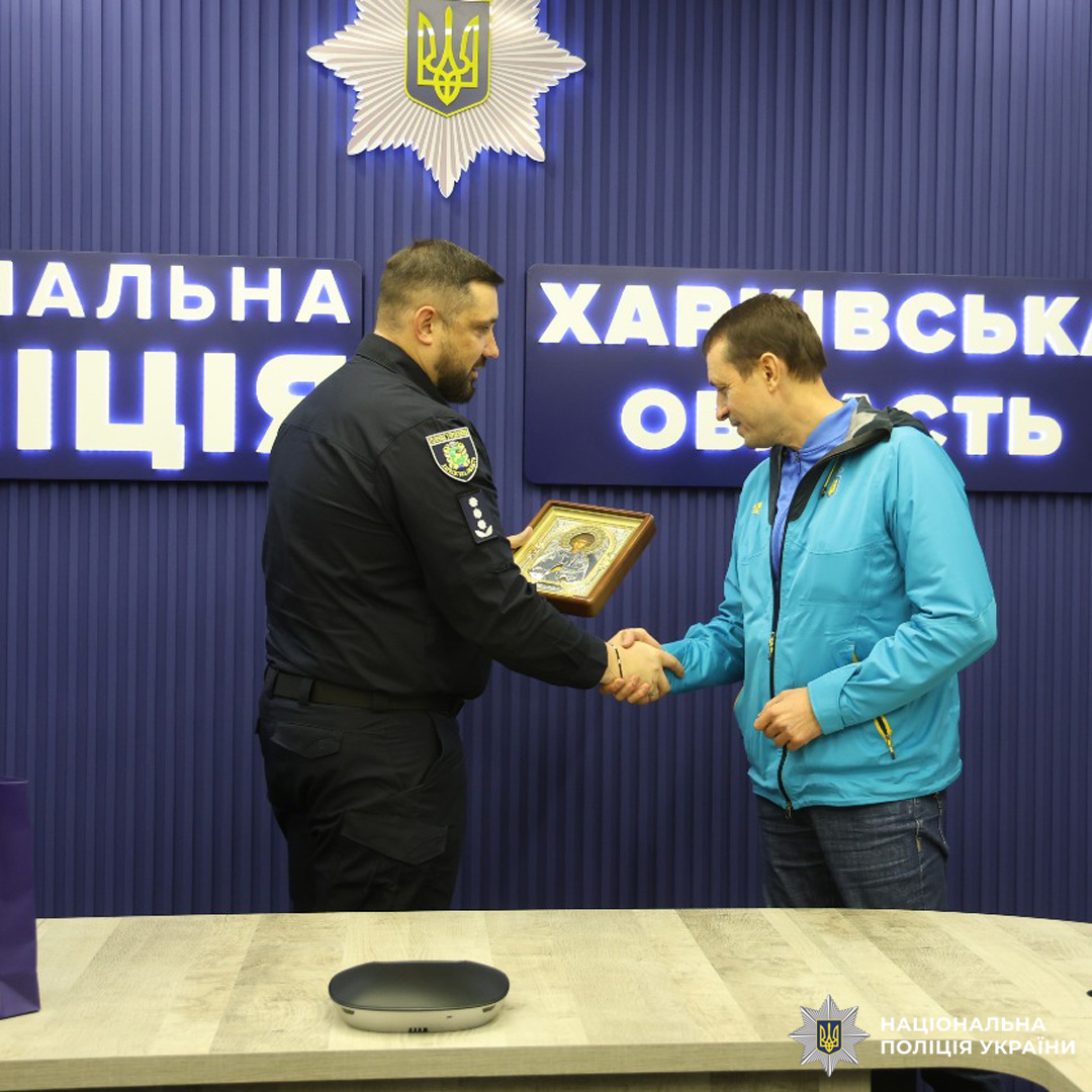 Поліцейський у формі передає ікону святого Пантелеймона чоловіку в синій куртці. Вони потискають руки на фоні банера з емблемою та написами «Національна поліція».