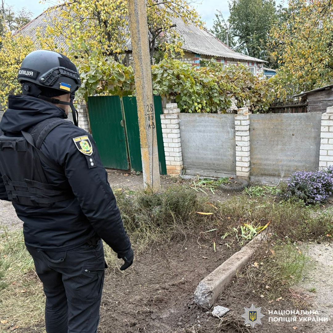 Поліцейський у чорній формі та шоломі з емблемою поліції стоїть на ґрунтовій дорозі біля житлової забудови, оглядаючи місце пошкодження. На землі зламаний електроопорний стовп, порушений ґрунт і рослинність.Біла цегляна стіна та частково видимий будинок із сірим дахом. Поруч рослинні уламки та кущ із фіолетовими квітами.