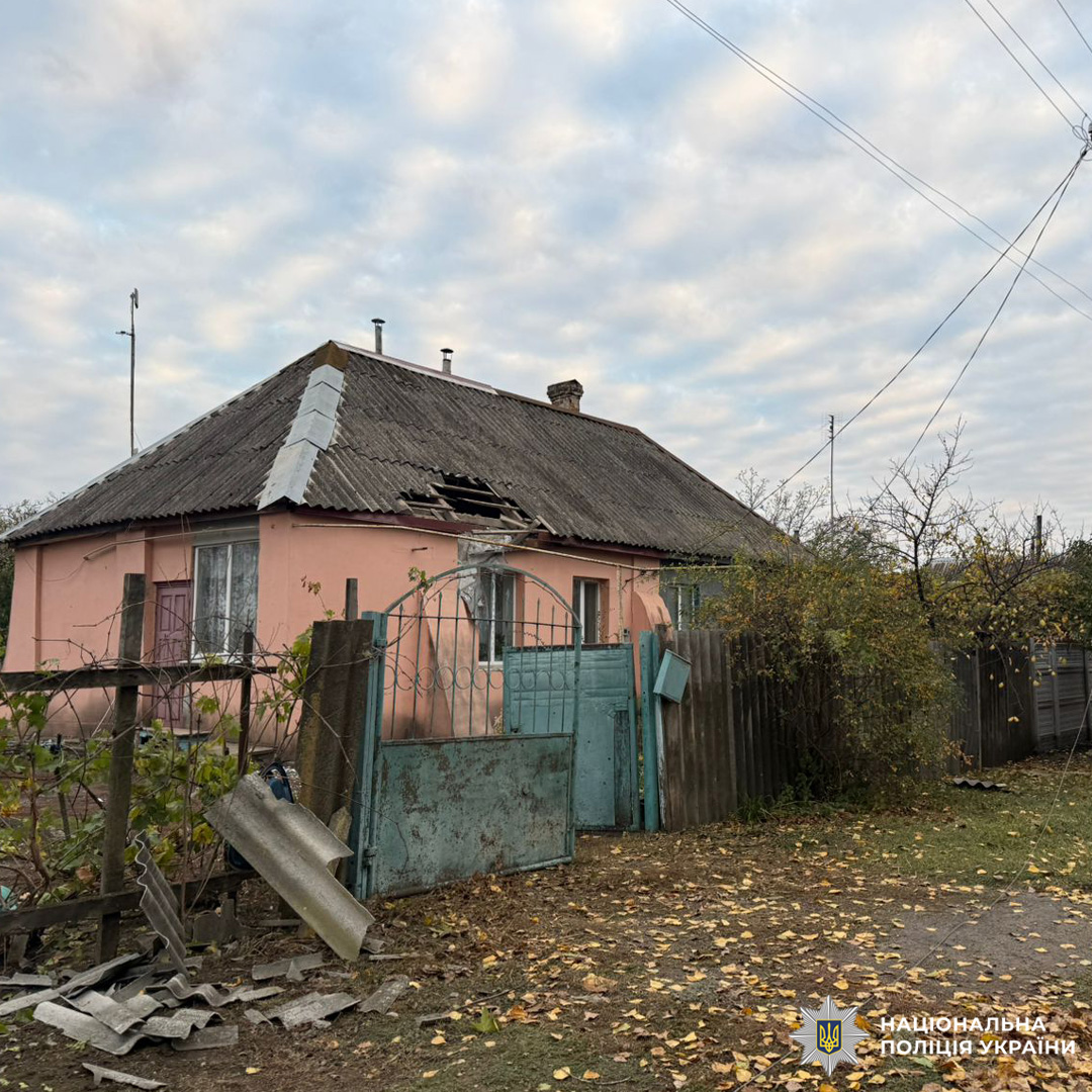 Одноповерховий житловий будинок рожевого кольору з пошкодженим дахом: видно відсутні черепиці або листи покриття. Перед будинком металева брама та паркан, частково відкриті й з ознаками зношеності.
