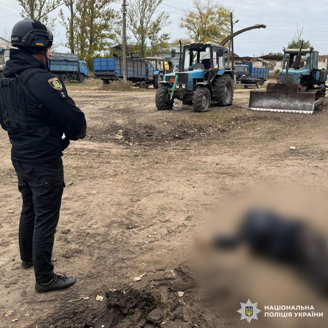 Офіцер у чорній формі та шоломі стоїть на відкритій ділянці з ґрунтовим покриттям.Місце події оточене синіми та зеленими тракторами й вантажівками, що вказує на сільськогосподарське або промислове середовище. На фоні дерева та будівлі.