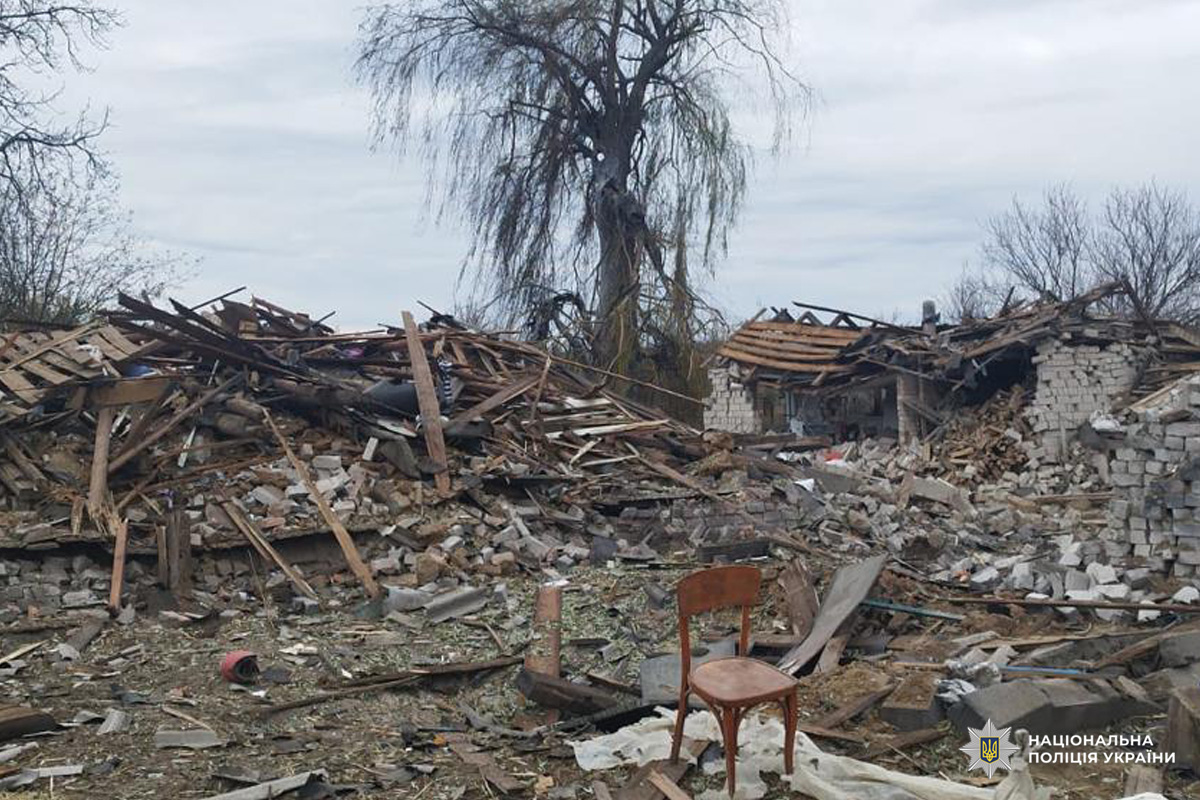 Зруйновані будівлі з обваленими дахами та стінами. На передньому плані дерев’яний стілець серед уламків. Видимі сліди вибуху або обстрілу.