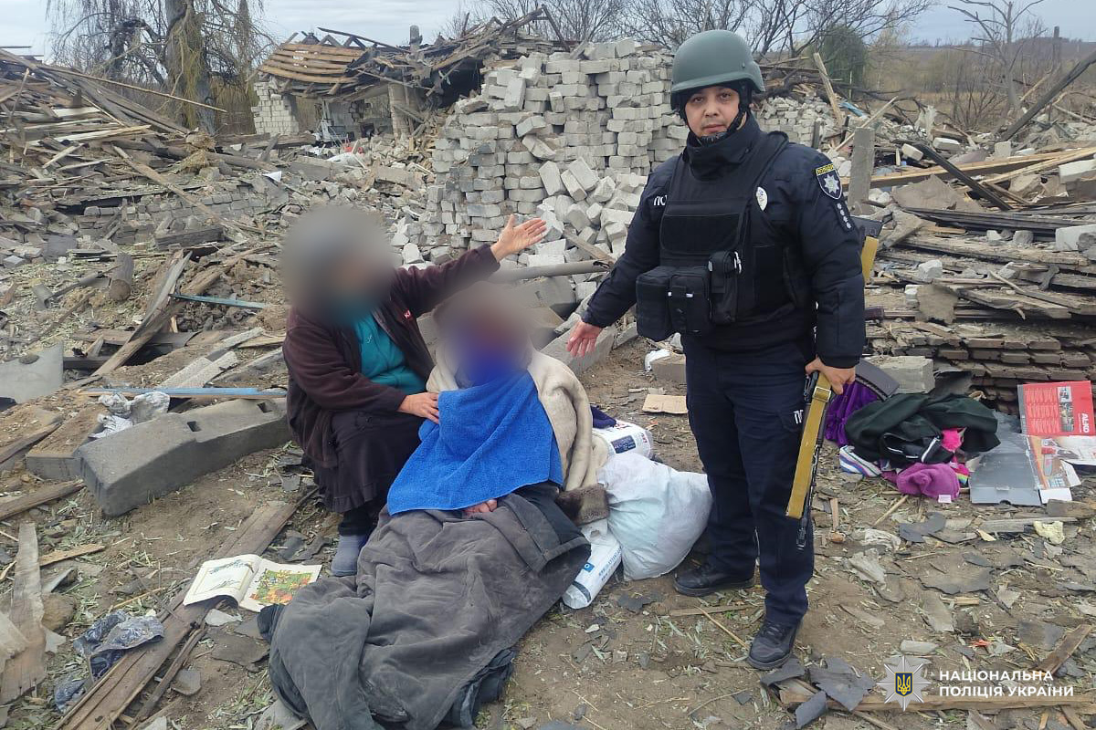 Поліцейський у шоломі та бронежилеті надає допомогу двом постраждалим, які сидять серед руїн, загорнуті в ковдри. На фоні зруйновані будівлі, уламки, особисті речі.