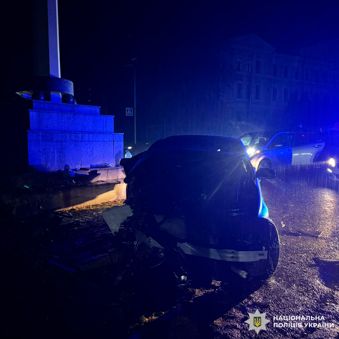 Пошкоджений автомобіль після зіткнення з кам’яним постаментом, поруч працює поліція, нічна зйомка під дощем.
