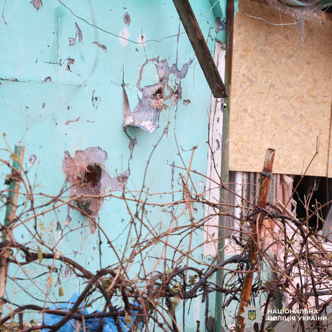 Пошкоджена стіна будівлі з численними отворами та тріщинами, ймовірно від уламків. Частково вікно закрито фанерою.