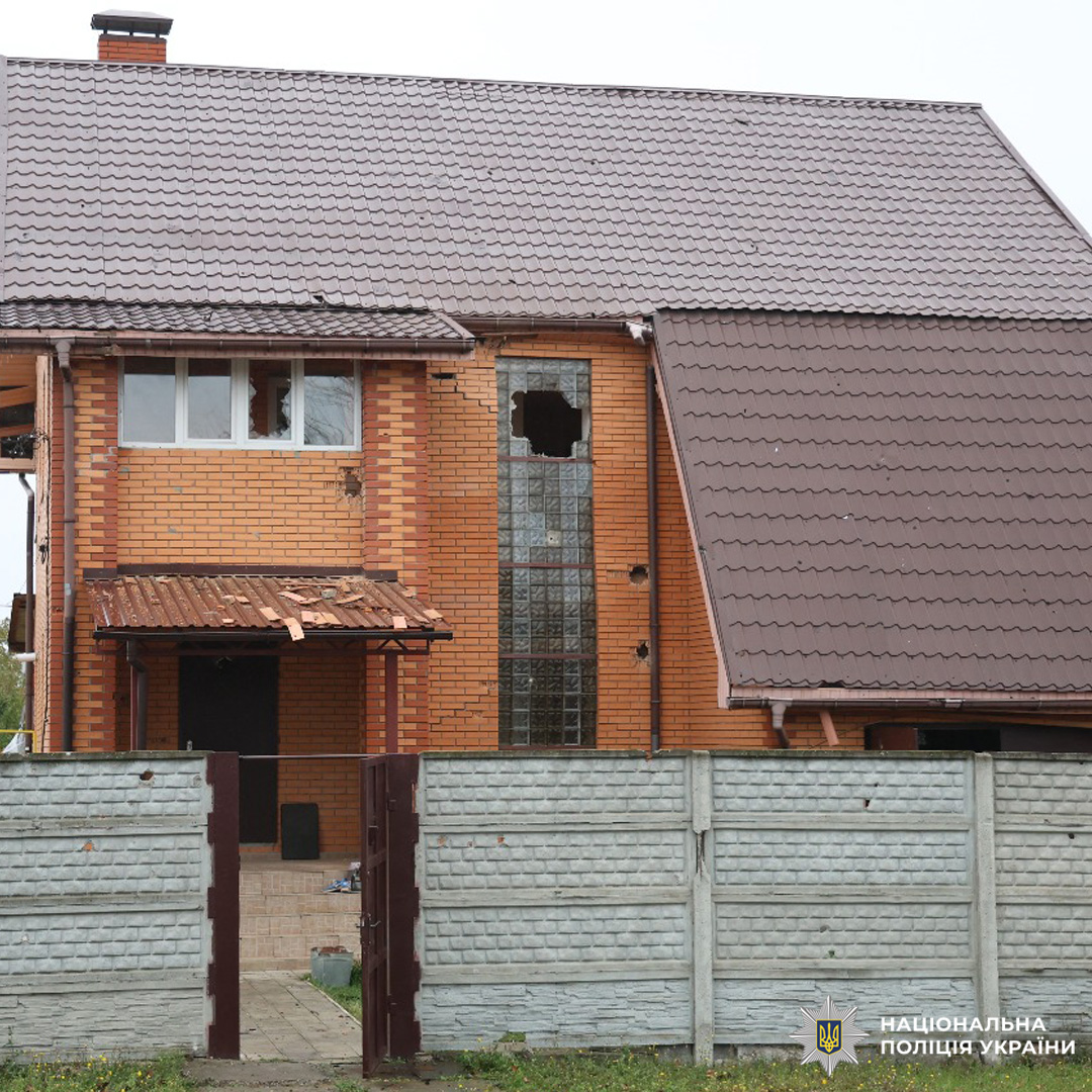 Цегляний будинок із коричневим дахом, пошкоджене скляне блокове вікно та навіс над входом. Сірий бетонний паркан, металева брама.