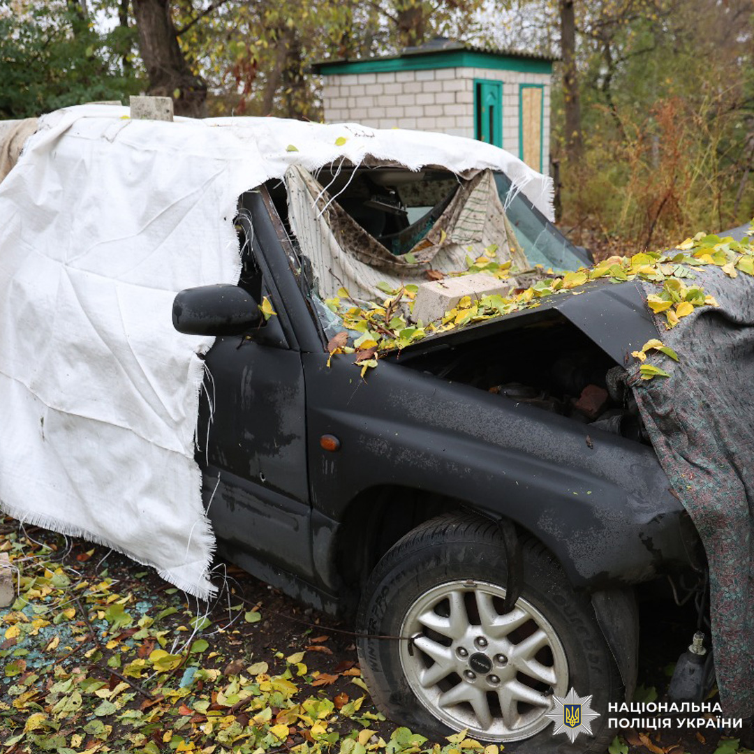 Пошкоджений чорний автомобіль із розбитим лобовим склом, деформованим дахом і капотом, частково накритий білим і сірим тентом. На тлі цегляна споруда з зеленою дверима.