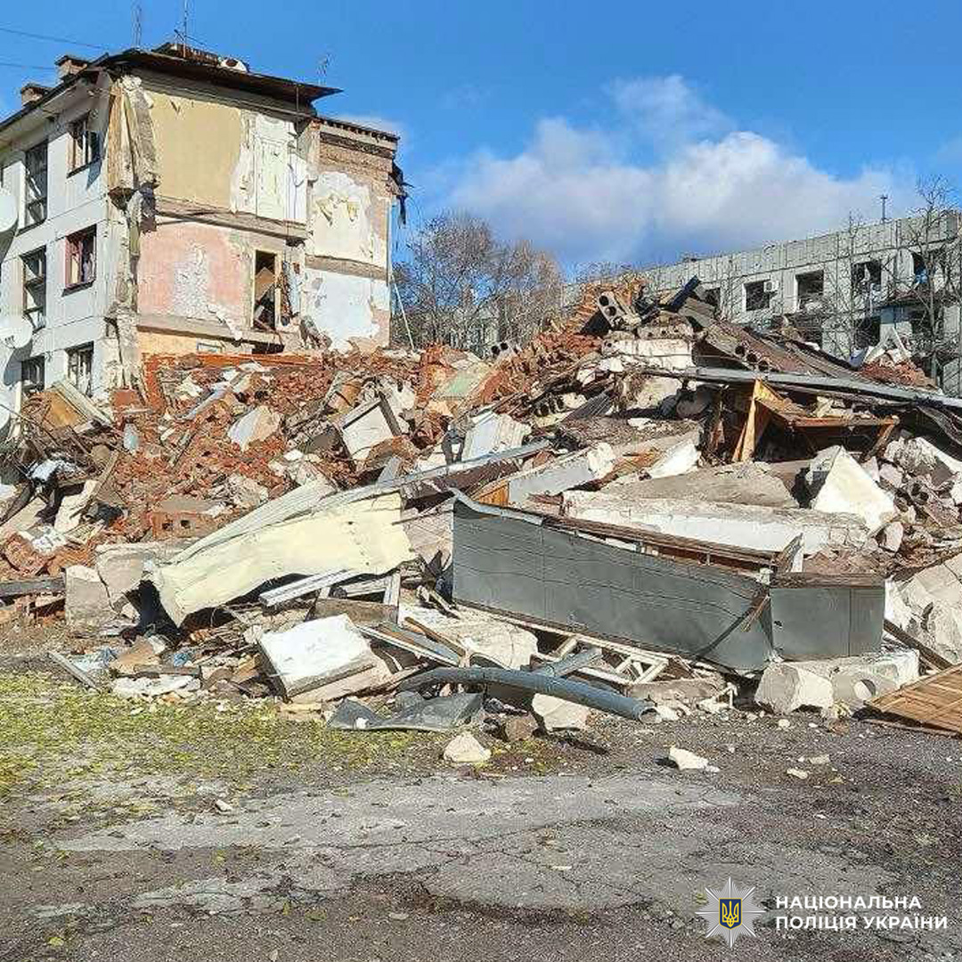 Частково зруйнована багатоповерхова будівля. На передньому плані велика купа уламків: бетон, цегла, скручений метал. У фоні ще одна пошкоджена споруда, дерева та ясне небо.