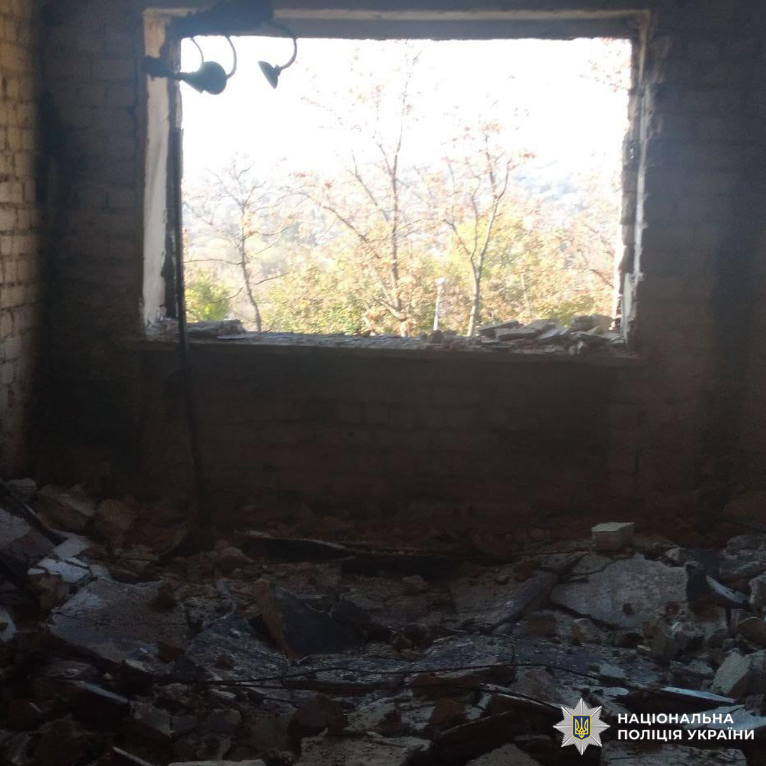 Інтер’єр зруйнованої кімнати з оголеною цегляною кладкою, вибитим вікном і розкиданими уламками. На стелі пошкоджений світильник. За вікном видно дерева та денне світло.