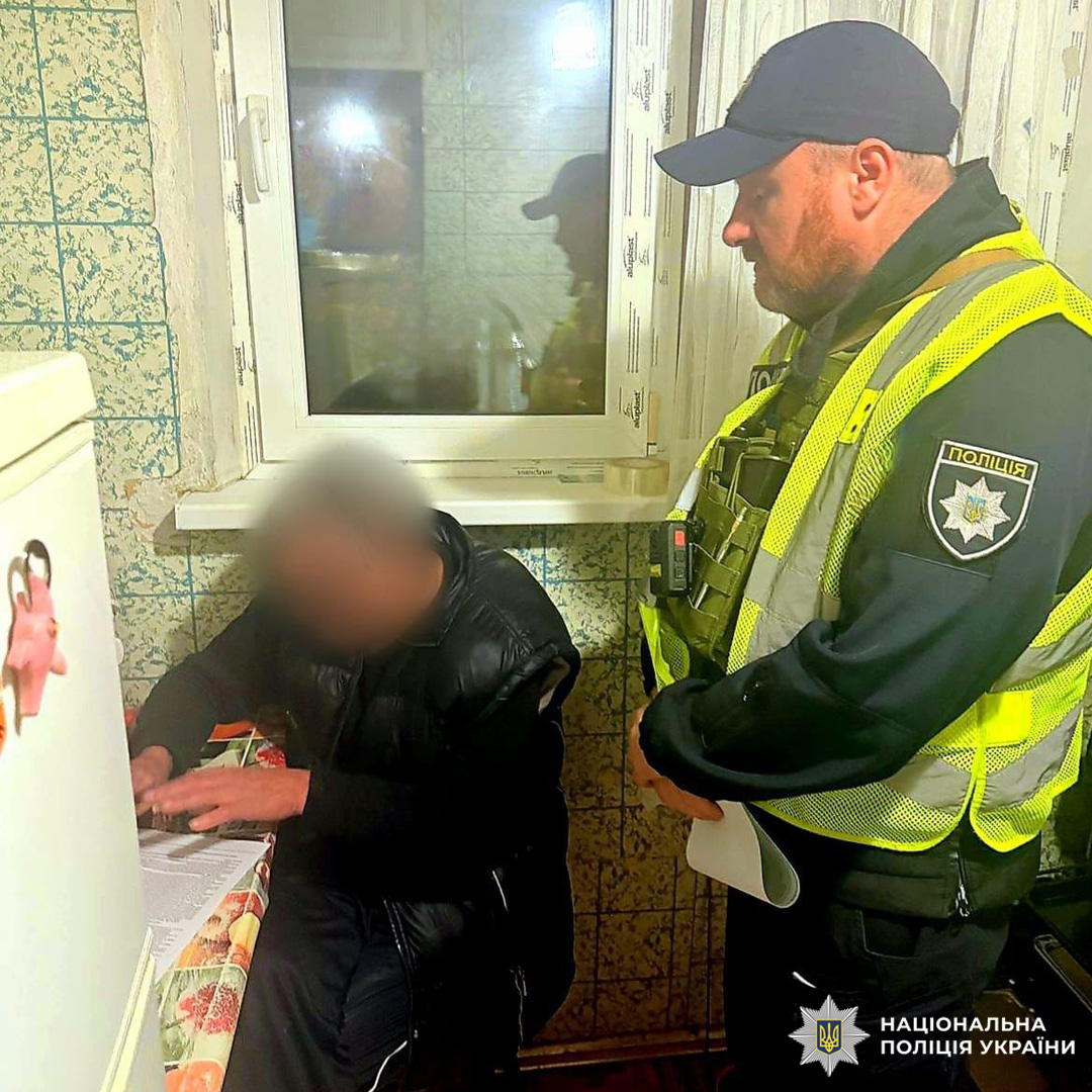 У домашньому приміщенні співробітник Національної поліції України у формі з високою видимістю спостерігає за громадянином, який сидить за столом і підписує документи. Обличчя цивільної особи розмито з міркувань конфіденційності.