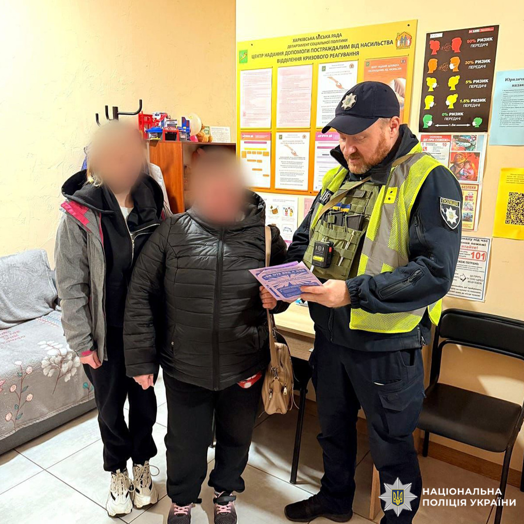 Співробітник Національної поліції України у формі з нагрудною камерою та світловідбивним жилетом проводить роз’яснювальну бесіду з двома громадянами. Обличчя цивільних осіб розмито з міркувань конфіденційності. На стіні плакати з інформацією про запобігання домашньому насильству.