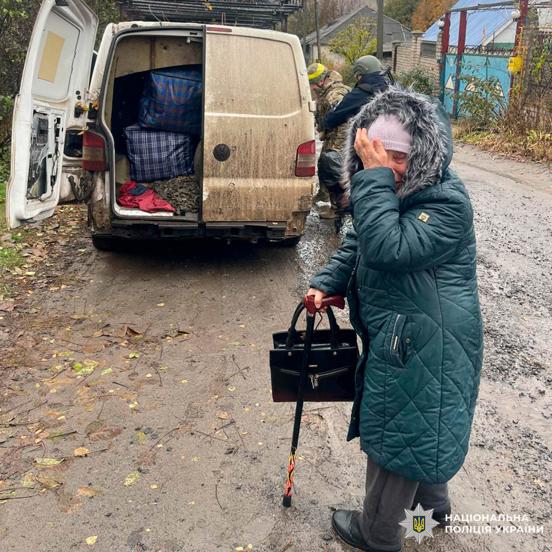 Сільська дорога з багнюкою, біля якої припаркований білий фургон з відкритими дверима. У фургоні видно великі сумки та контейнери. На передньому плані літня жінка в зеленому стьобаному пальті з хутряним капюшоном, яка тримає палицю та чорну сумку, прикриваючи обличчя рукою.