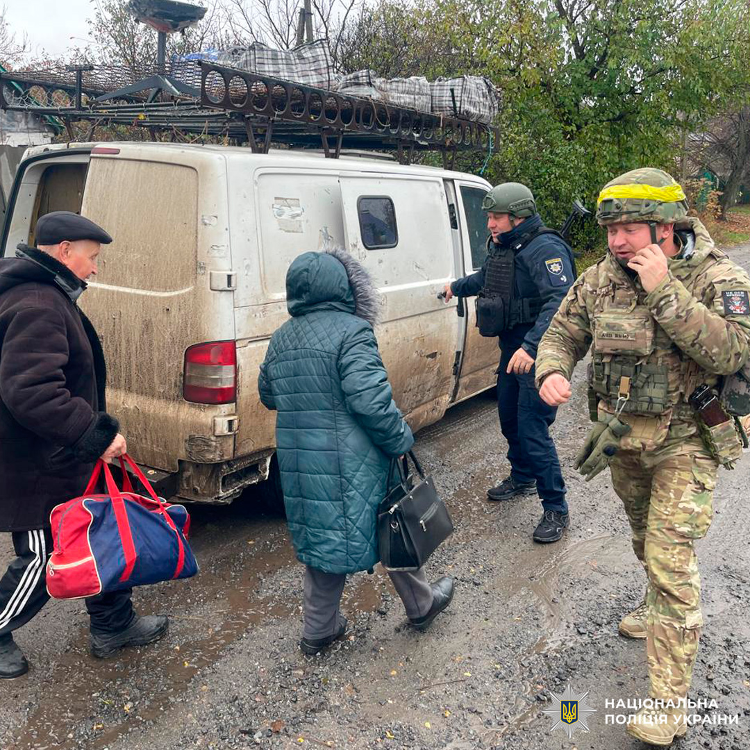 Фургон забруднений, на даху закріплені матраци та металеві конструкції. Один поліцейський у камуфляжі та шоломі з жовтою стрічкою говорить по телефону, ймовірно координуючи дії. Другий поліцейський допомагає жінці в синьому пальті з сумкою, цивільний чоловік у чорному пальті несе спортивну сумку.