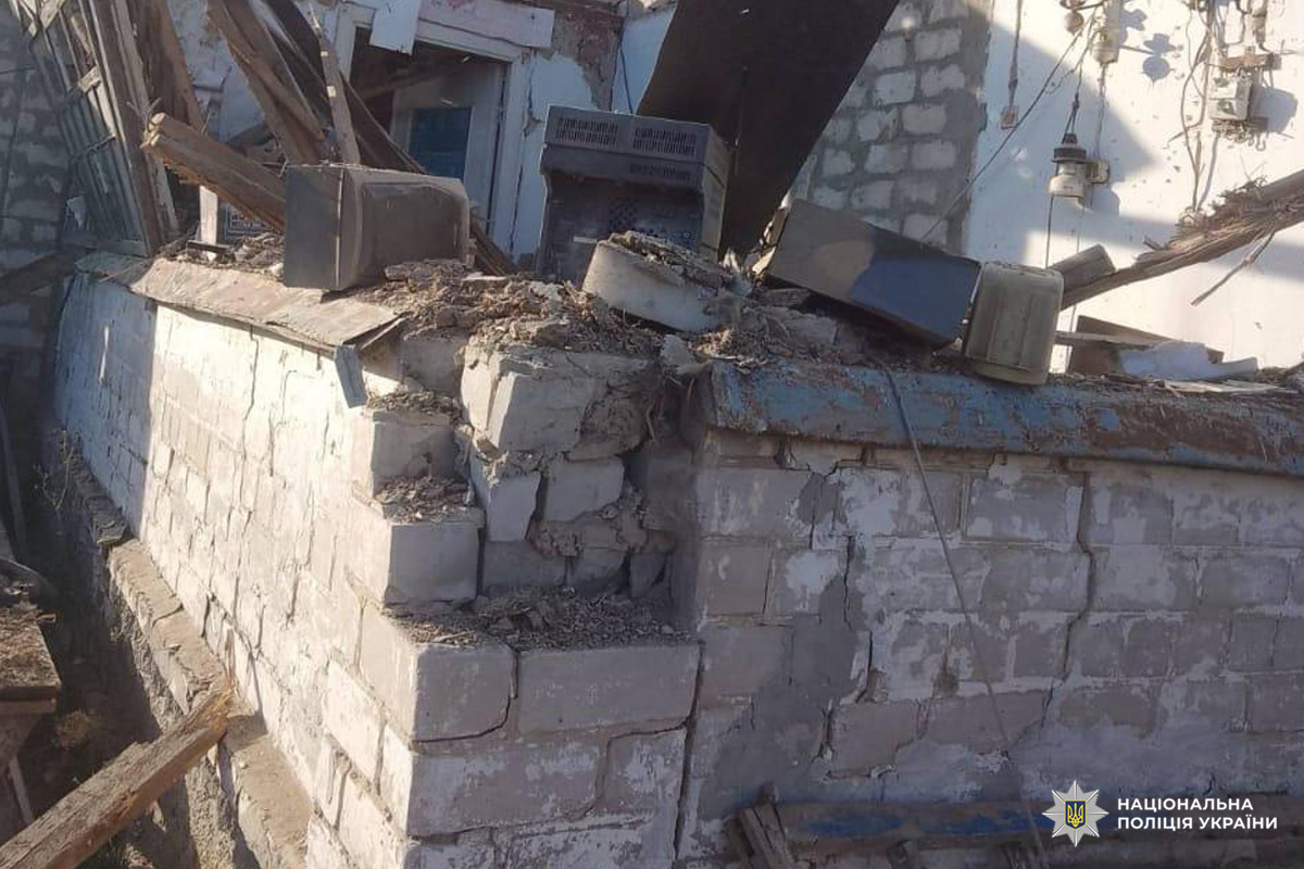 На фото частково обвалена стіна з бетонних блоків, значні тріщини та відсутні фрагменти конструкції. На уламках побутова техніка, дерев’яні балки, металеві листи та інші предмети домашнього вжитку.