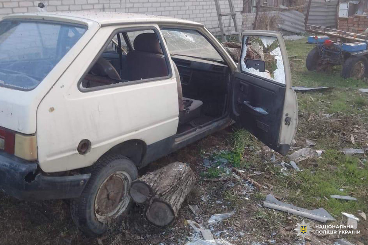 Авто стоїть на ділянці біля цегляної будівлі, двері водія відчинені, салон частково розібраний. Під задньою частиною дерев’яні колоди, ймовірно, для підтримки. На задньому плані трактор та будівельні матеріали.