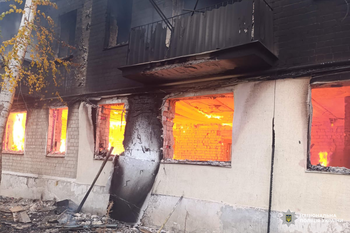 На фото багатоповерхова споруда, охоплена вогнем: полум’я видно крізь кілька вікон першого поверху, дах і фасад частково пошкоджені. На землі уламки конструкцій.