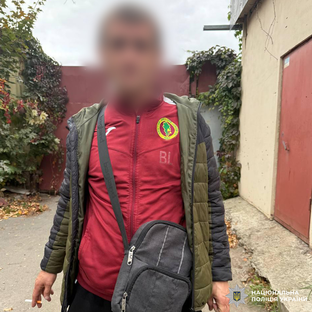 Чоловік перед червоними воротами та бетонною стіною. Одягнений у червону кофту з логотипом і літерами «BI» на грудях, поверх темно-зелена куртка та чорна сумка через плече.